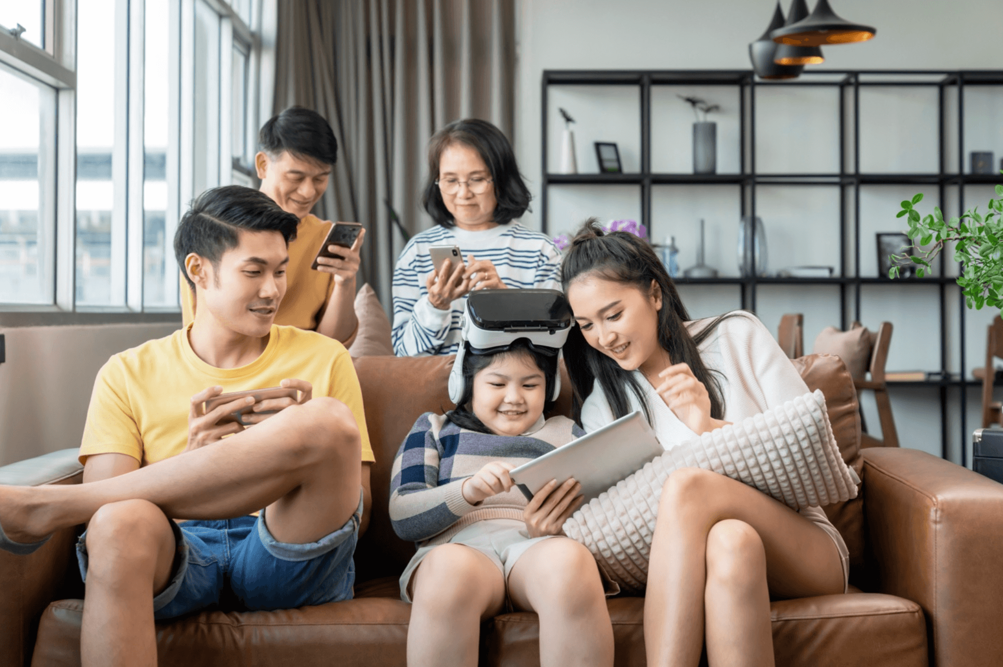 Family congregates in the living room using a variety of smart devices like a VR headset, tablet, and smart phones