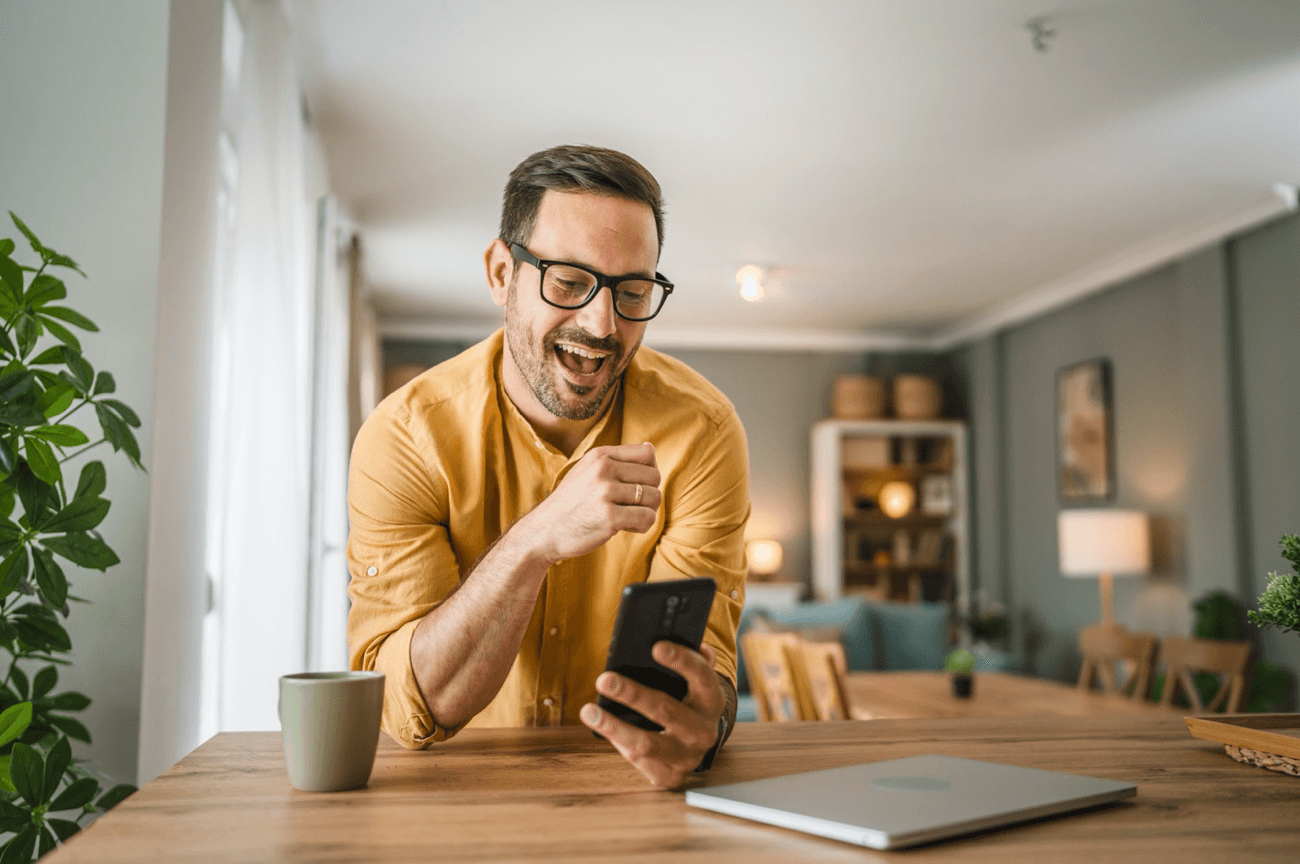Happy man looks at phone while sitting at a table with a coffee mug and laptop