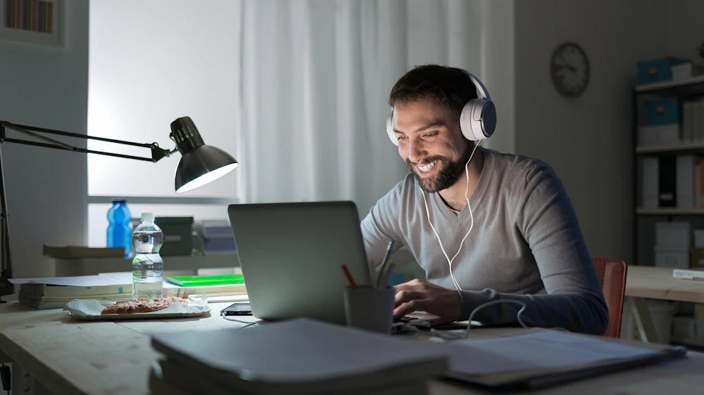 Man with headset uses laptop at night illuminated by desk lamp