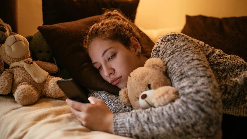 Young woman lies on bed clutching teddy bear while looking at her smartphone