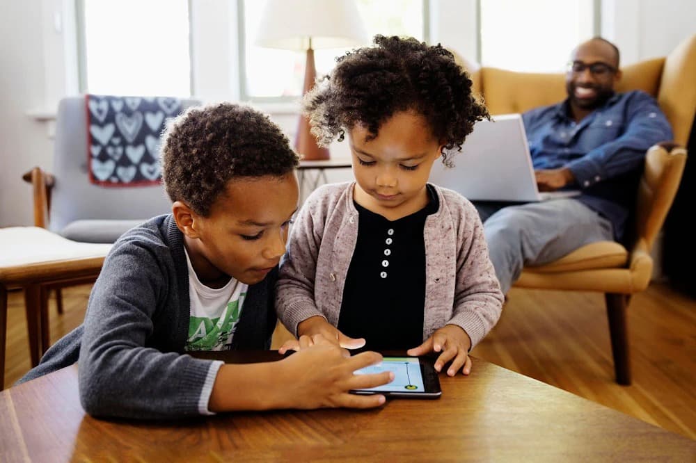 Children share a tablet while father uses a laptop and looks on
