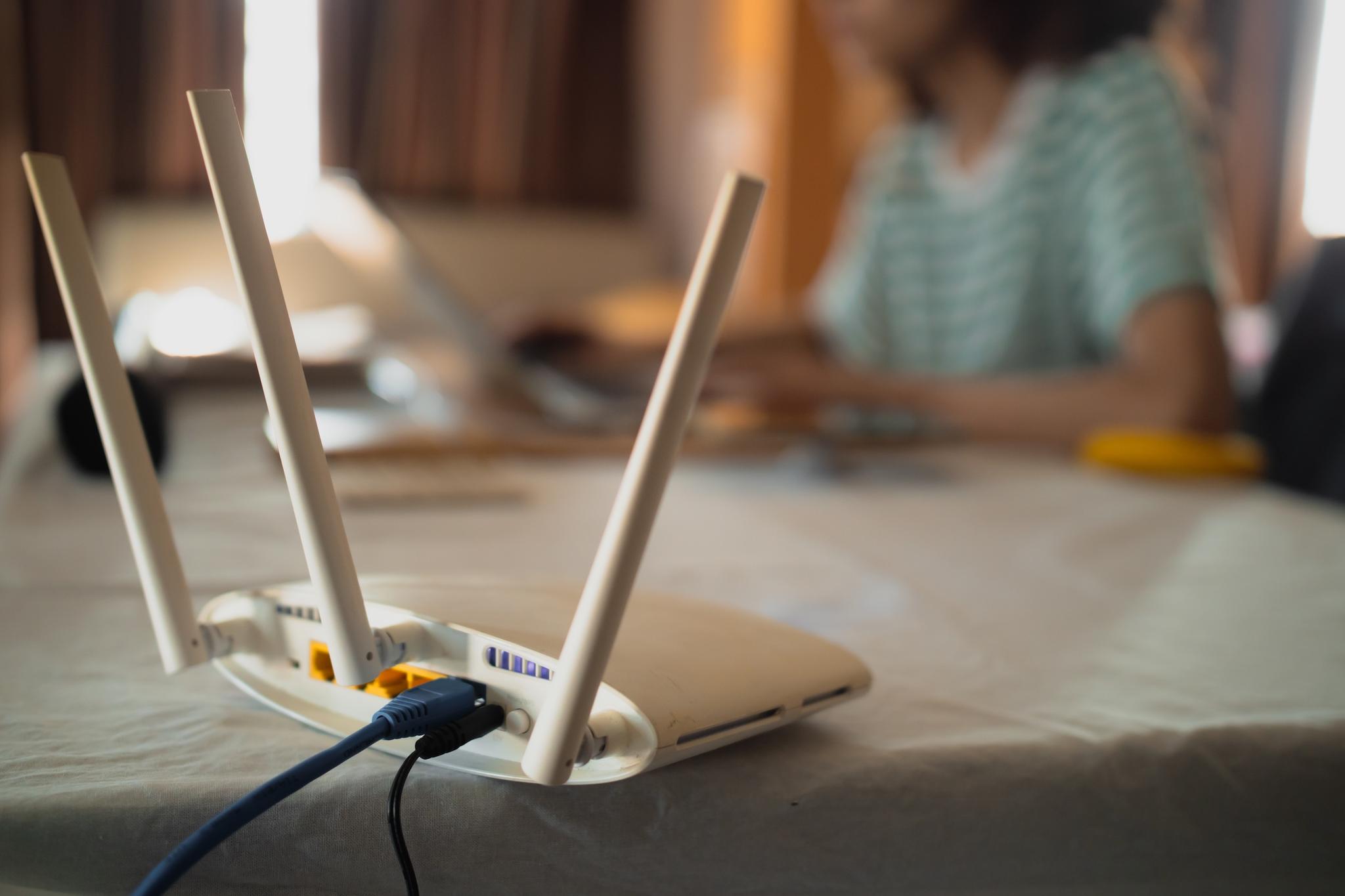 A Router in the foreground with cables plugged into it while person works on a laptop in the background