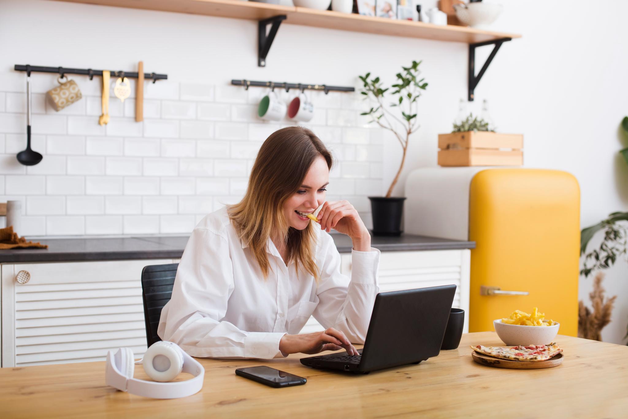 Young woman working on a laptop while eating in the kitchen.