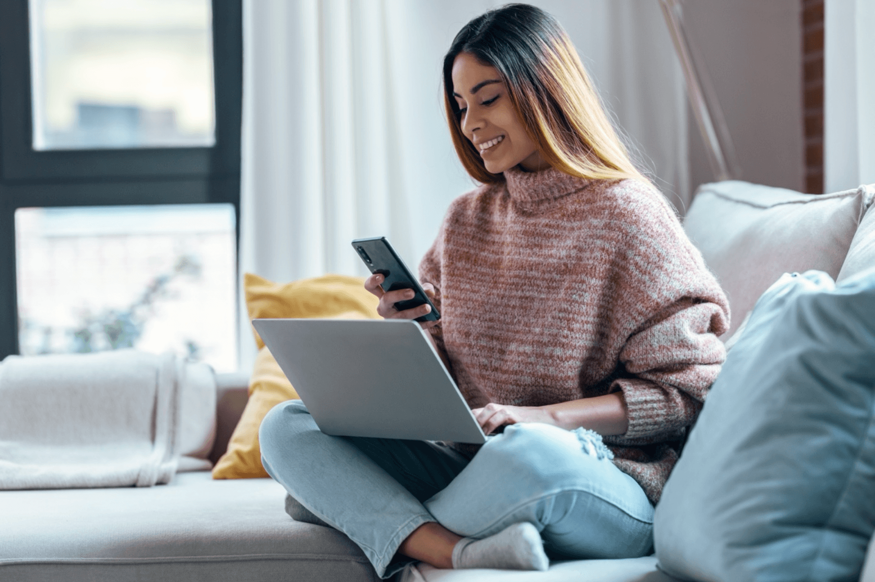 Young woman sits on couch with laptop and phone in hand