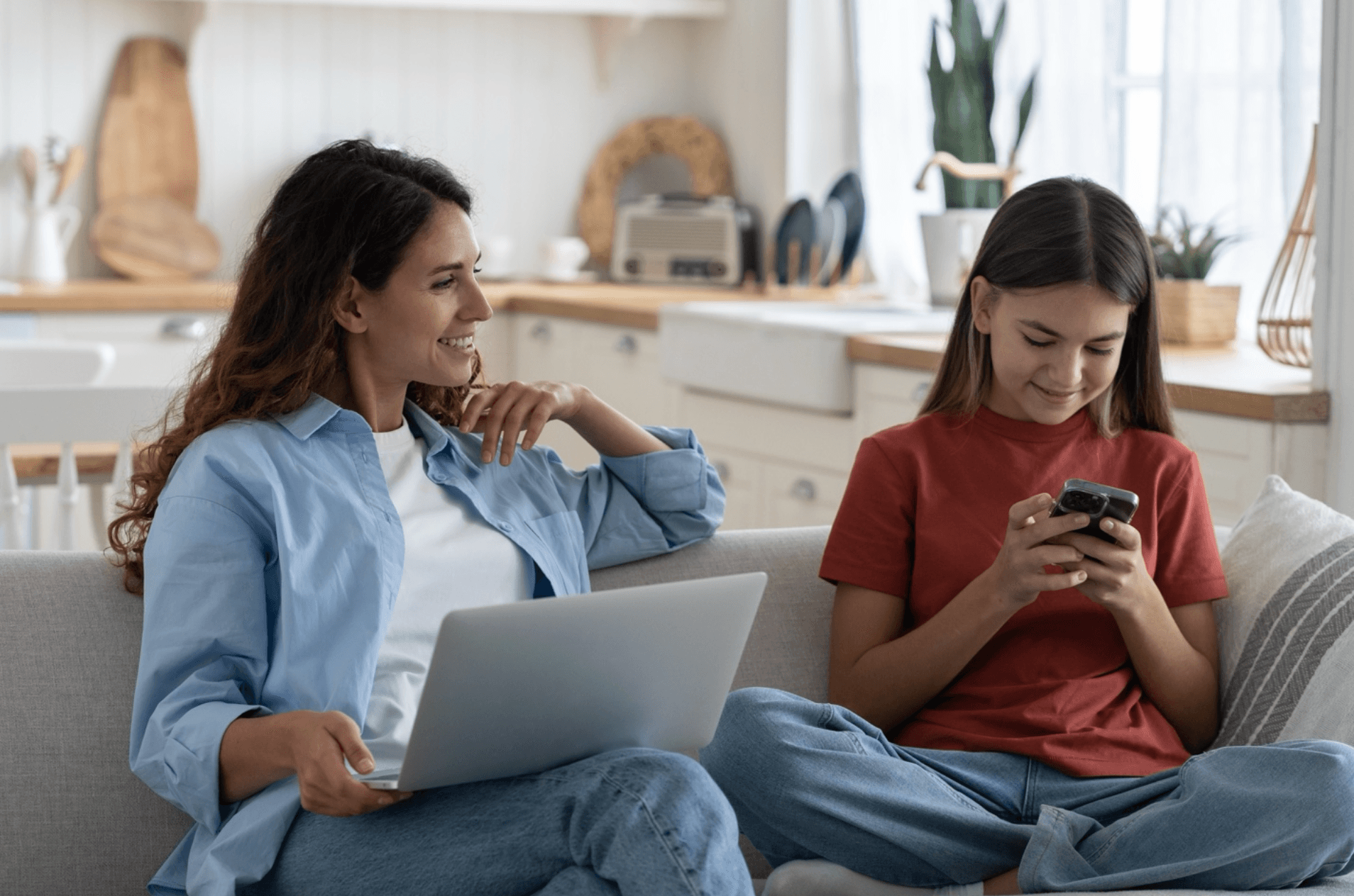 Smiling mother with laptop on her lap looks at her happy daughter using a smartphone