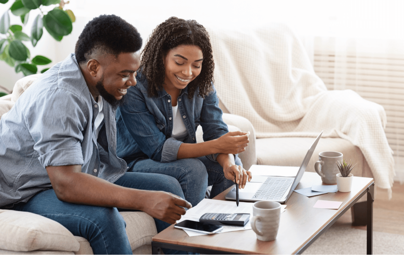 Husband and wife sit on couch to budget with calculator and laptop