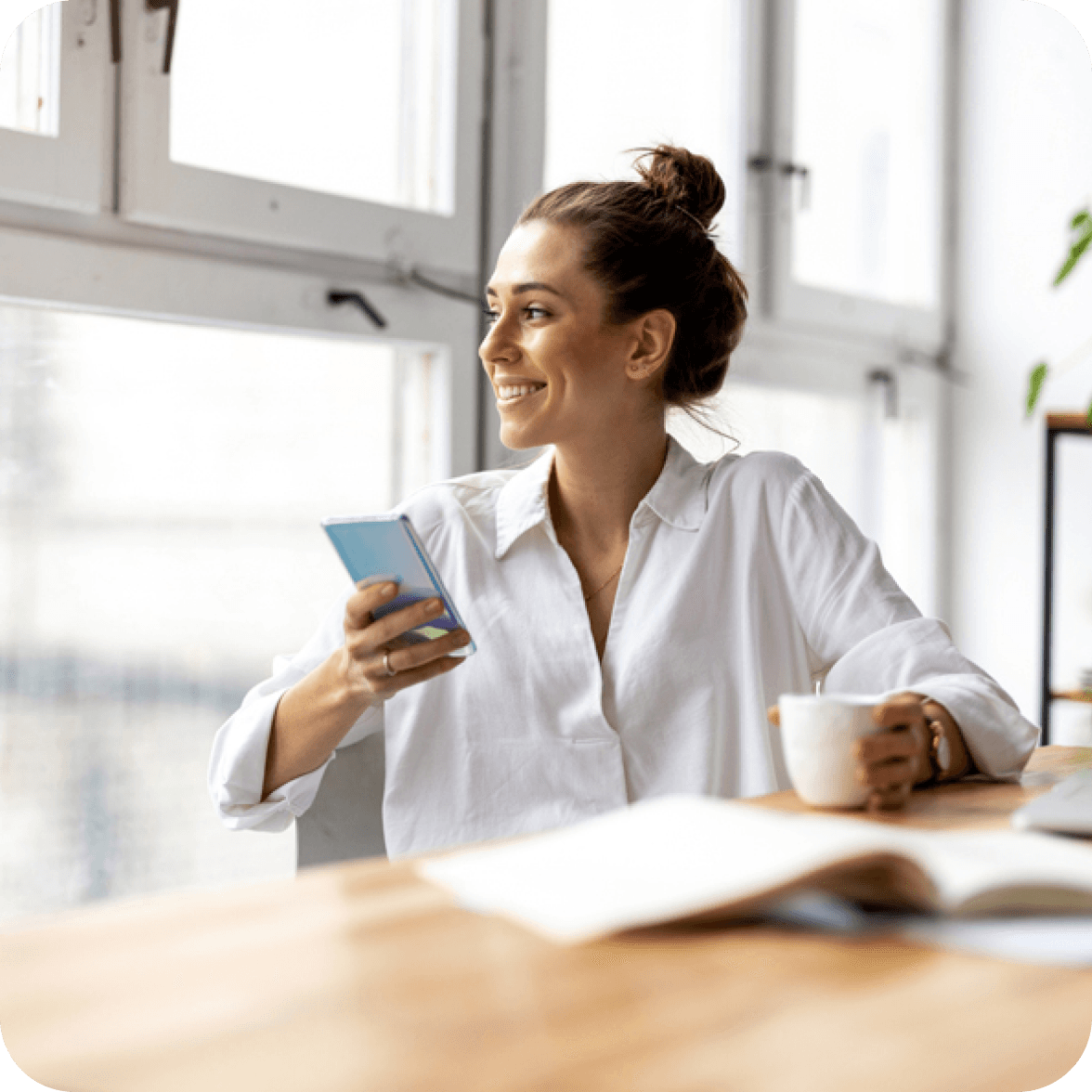 Woman sitting at a desk with phone in one hand, coffee cup in the other, looking out a window.