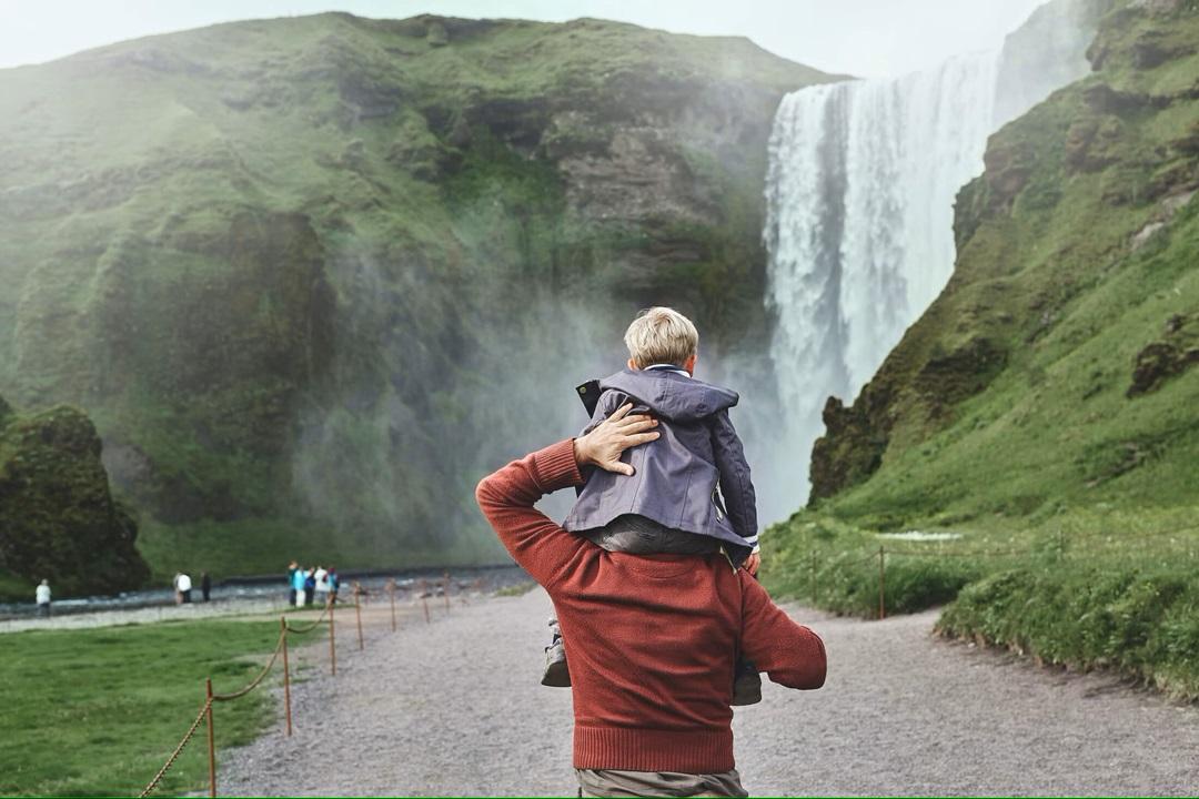 Son on dad's shoulders walking towards waterfall in Iceland.