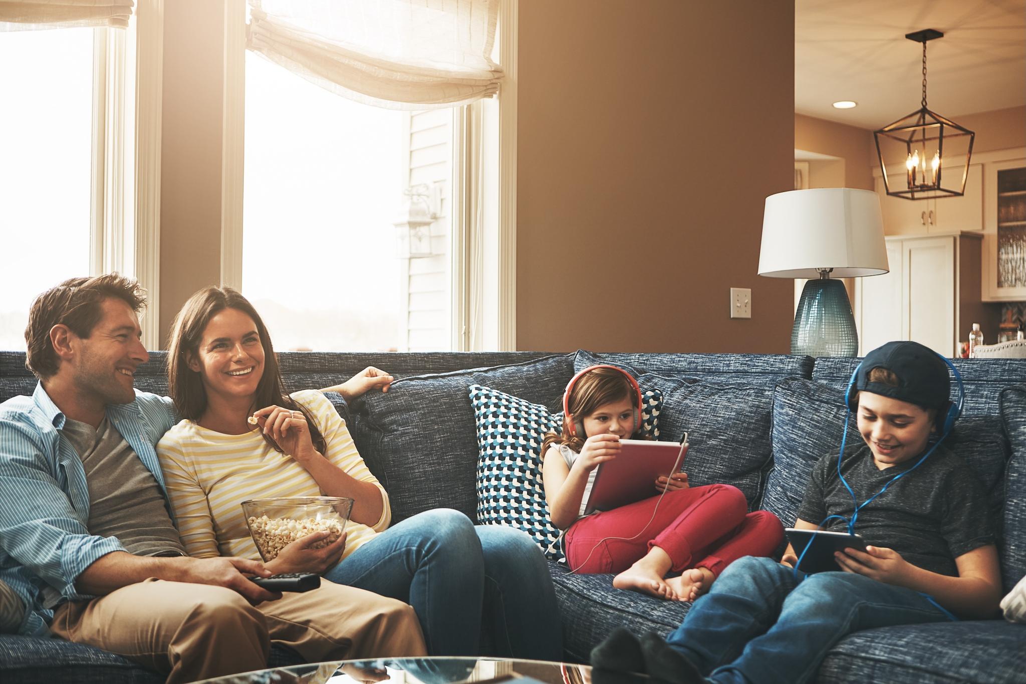 Parents watching a movie and kids playing on tablets sit on a couch together