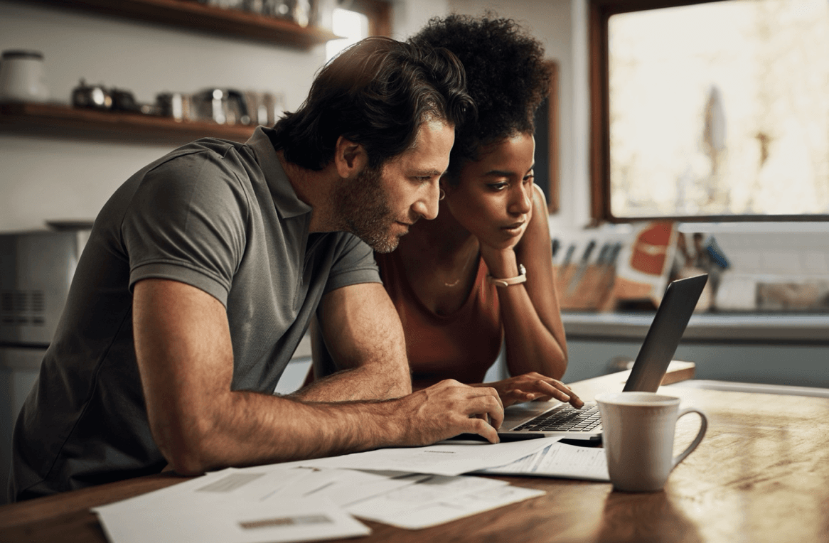 Man and woman look intently at laptop with papers scattered around a table