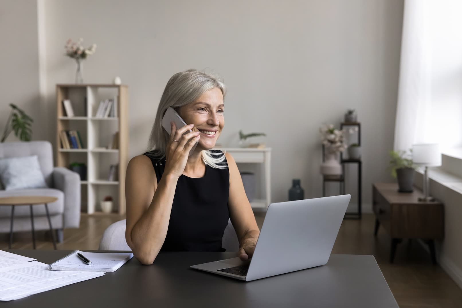 Smiling woman sitting at a desk, working on a laptop in her home office, making mobile phone calls.
