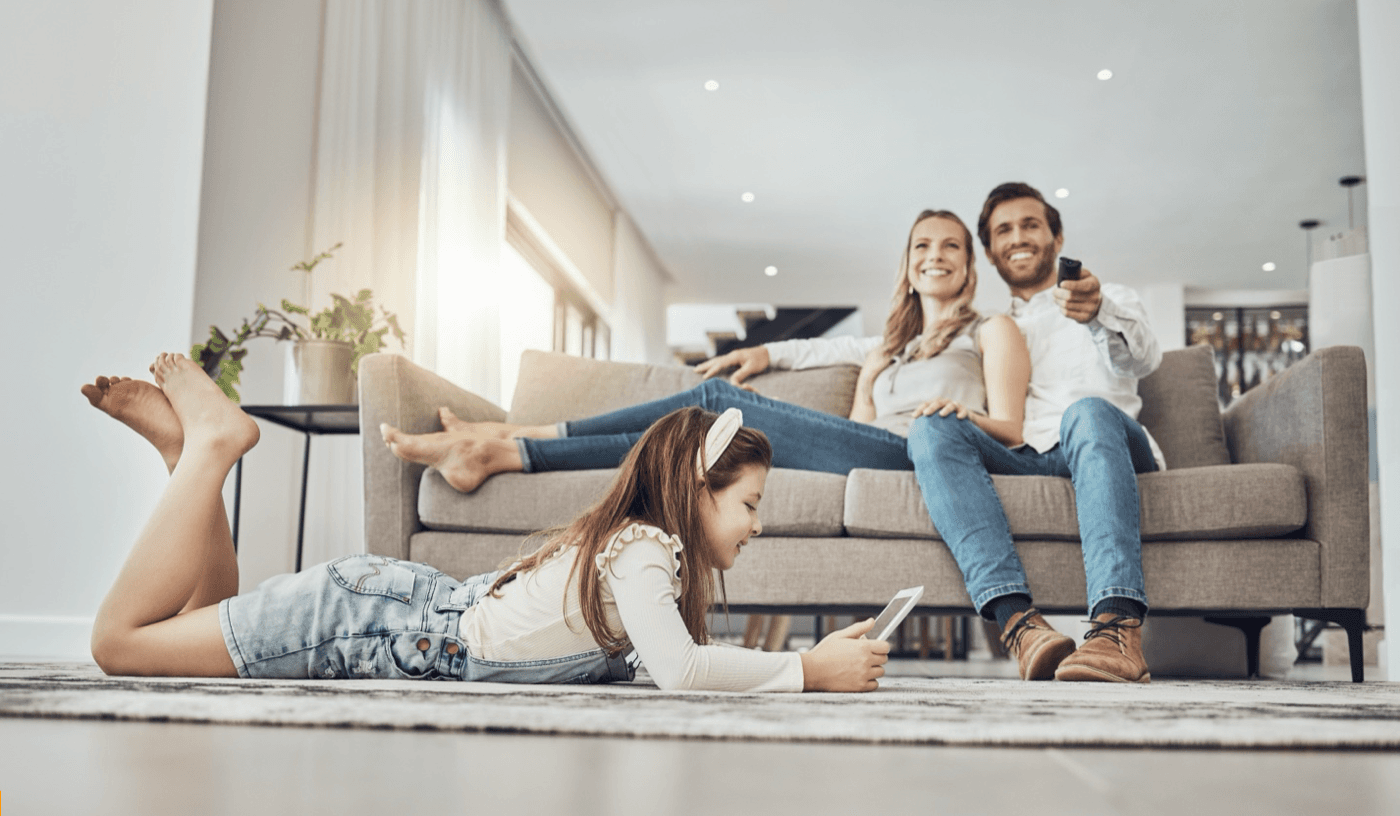 Parents sit on couch with remote while daughter watches something on her tablet on the floor